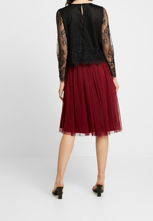 Musta pitsinen pitkähiha toppi, jossa on avainreikäselkä, yhdistettynä burgundinväriseen tüllihameeseen. Kirkkaat korot täydentävät asukokonaisuuden.