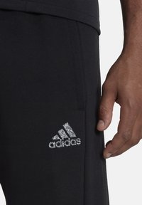 Svarta träningsbyxor med en mjuk textur, med en silverreflekterande Adidas-logotyp på vänster sida och en sidoficka.