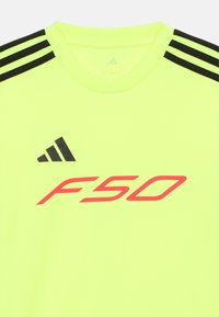 Keltainen toiminnallinen urheilupaita, jossa mustat kolmisuoraista raitaa olkapäillä ja edessä rohkea punainen "F50" -logo. Sileä pinta, pyöreä kaulus.