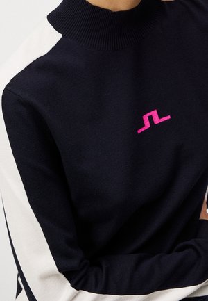 Navy trui met witte strepen op de mouwen en een klein roze abstract logo op de borst, gedragen door een persoon met gekruiste armen.