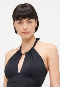 Fato de banho preto com alças halter em tecido liso apresenta um design em forma de buraco na zona do peito e detalhes em dourado na gola.