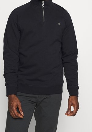 Mężczyzna ubrany w czarny pulower z długim rękawem, z ćwiartkowym zamkiem błyskawicznym i małym złotym haftowanym logo na lewej klatce piersiowej, w połączeniu z ciemnymi spodniami.