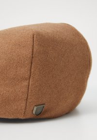 Gorra de fieltro marrón con forma redondeada, cuenta con un pequeño logotipo de metal embellecido y una textura suave. Detalles de costura visibles.