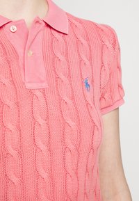 Różowa dzianinowa koszulka polo z warkoczowym wzorem, krótkimi rękawami i klasycznym kołnierzykiem. Niebieskie logo na lewej piersi.