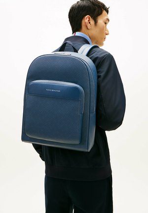 Giovane uomo con giacca blu navy che porta uno zaino Tommy Hilfiger blu testurizzato, su uno sfondo chiaro uniforme, di profilo.