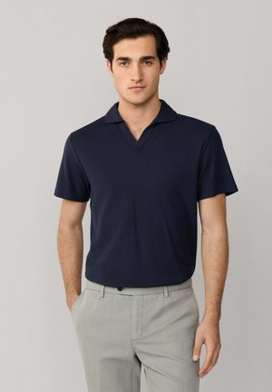 Jonge man met donker haar, gekleed in een marineblauw overhemd met korte mouwen en een kraag, en lichtgrijze broek, staand tegen een effen achtergrond.