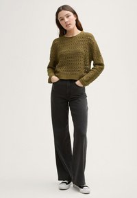 Olijfgroen gebreid sweater met open haakpatroon, gecombineerd met zwarte high-waisted wijde jeans en zwarte sneakers met witte zolen.