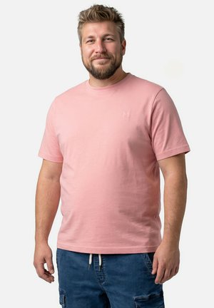 Bärtiger Mann mit kurzen Haaren, der ein hellrosa kurzärmeliges T-Shirt und blaue Cargo-Shorts trägt, steht vor einem schlichten hellen Hintergrund.