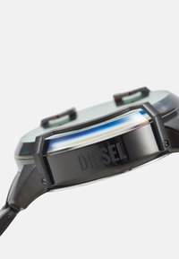 Schwarze metallische Uhr mit einem runden Gehäuse, ausgestattet mit einer schillernden Lünette und dem eingravierten Schriftzug "DIESEL" an der Seite. Das Glas ist leicht gewölbt.