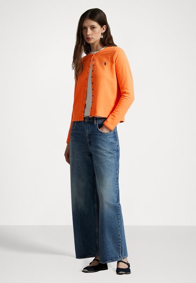 Orange knit cardigan with round neckline, button front, worn over a white top. Paired with high-waisted blue wide-leg jeans and black flats.