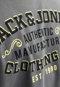 Grijze T-shirt met opvallende zwart en gele grafische tekst: "Jack & Jones Authentic Manufacturing Clothing EST. 1990," met decoratieve accenten.