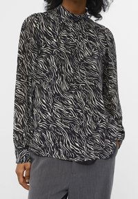 Blouse à manches longues en tissu noir et crème avec un imprimé zèbre ondulé. Dotée d'un col montant et de détail froncé aux épaules.