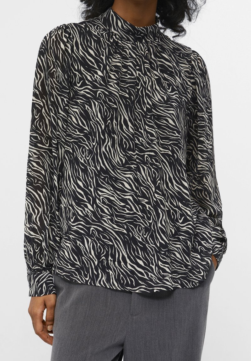 Blouse à manches longues en tissu noir et crème avec un imprimé zèbre ondulé. Dotée d'un col montant et de détail froncé aux épaules.