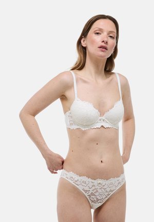 Femme portant un soutien-gorge en dentelle blanche et une culotte assortie en dentelle, debout avec une main ajustant la culotte sur un fond uni.