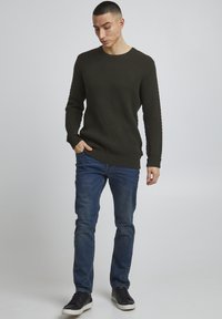 Ciemnozielony sweter o strukturze dzianiny z okrągłym dekoltem, w połączeniu z niebieskimi dżinsami i czarnymi trampkami. Prosty, codzienny design.