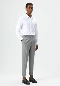 Witte blouse met lange mouwen en knopen, gecombineerd met op maat gemaakte grijze broek voorzien van een riem en manchetten. Zwarte loafers maken de look af.