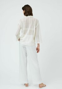 White, lightweight long-sleeve top with an open-knit design and round neckline, paired with loose-fitting, light-colored trousers.