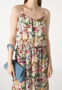 Femme portant une robe florale sans manches tenant un petit sac à main en denim bleu à ses côtés sur un fond uni.
