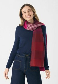 Geruite rode en roze sjaal met een zachte textuur, om de nek gedrapeerd, contrasterend met een blauwe gebreide trui, gecombineerd met donkerblauwe spijkerbroek.