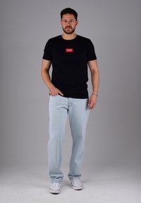 Mann mit Bart, der ein schwarzes T-Shirt mit rotem Logo, hellblaue Jeans und weiße Sneaker trägt und vor einem schlichten grauen Hintergrund steht.