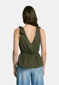 Blusa senza maniche di colore verde oliva con schiena a V, vita arricciata e dettagli decorativi a cravatta sulle spalle, abbinata a jeans azzurri chiari.