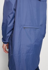 Vandtæt blå jakke med en glat tekstur, hætte og elastiske manchetter. Har en lynlåslomme på ryggen til opbevaring.