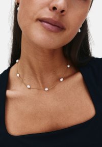 Guldhalsband med oregelbundet placerade vita pärlor, med en len yta och en delikat design, bärs med en svart topp.