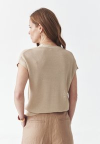 Top de punto beige con mangas cortas, que presenta una tela texturizada. Observado desde la parte trasera, combinado con pantalones cortos de lino marrones.