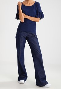 Donna che indossa un top in pizzo navy blu con maniche a gomito, jeans larghi scuri e sneaker bianche, in piedi con le braccia incrociate.