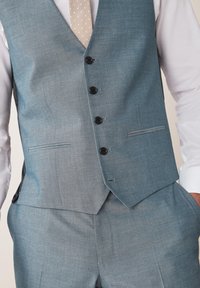 Hellblaues Anzugsweste mit strukturierter Oberfläche, fünf schwarzen Knöpfen und zwei Seitentaschen, getragen über einem weißen Hemd und einer beige gepunkteten Krawatte.