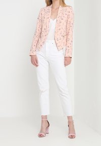 Femme debout portant un blazer floral rose clair, un haut blanc, un jean blanc aux ourlets effilochés et des talons ouverts roses à bloc.