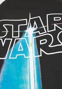 Musta t-paita, jossa on rohkea "STAR WARS" -teksti valkoisena, taustalla sinivalkoinen graafinen kuva valokeilasta ja tähtisateesta.