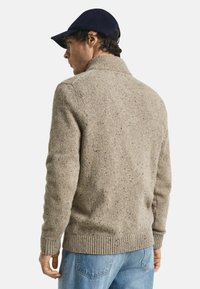 Mann trägt einen beige gesprenkelten Strickpullover, hellblaue Jeans und eine marineblaue Mütze, seitlich nach links gewandt, mit sichtbarem Rücken und Seite vor weißem Hintergrund.