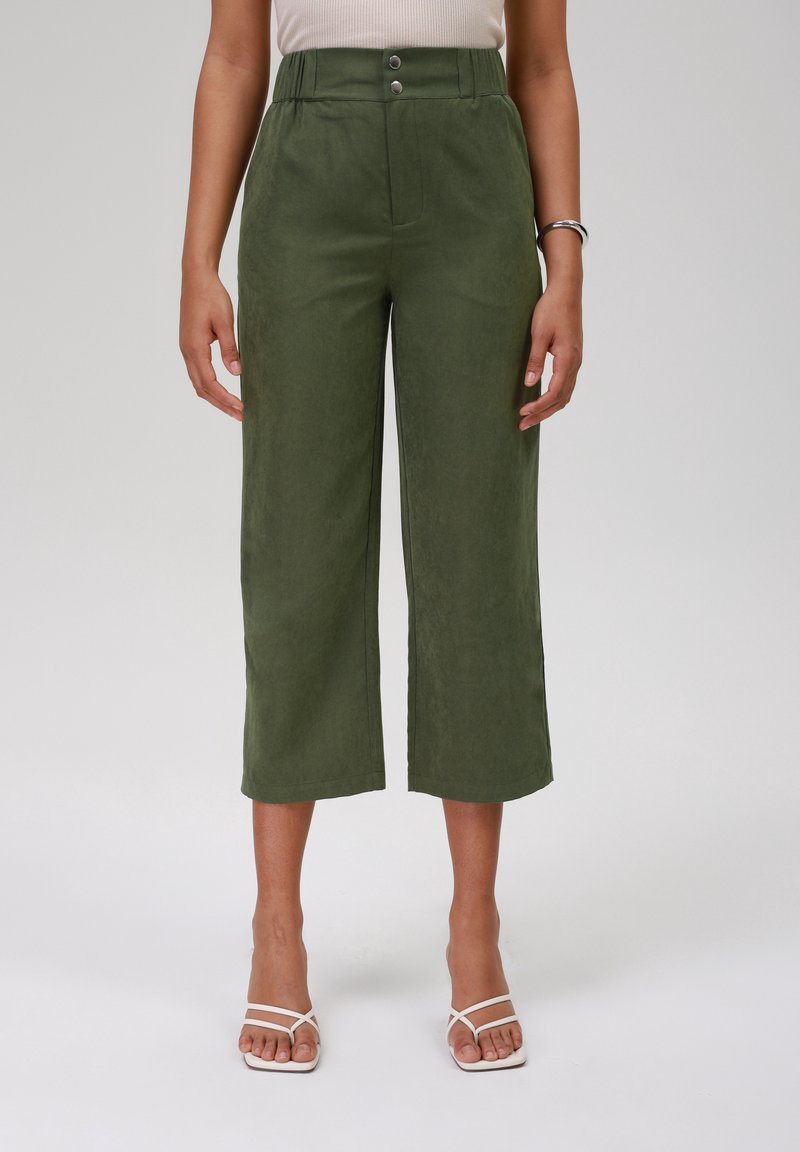 Femme portant un pantalon court à taille haute, jambes larges, couleur vert olive, avec fermeture à double bouton, et des sandales blanches à talons avec des lanières.
