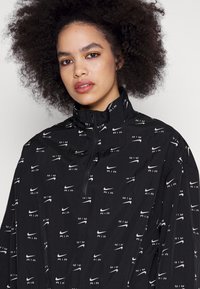 Jachetă neagră tip windbreaker, cu guler înalt, închidere cu fermoar pe jumătate și un model repetat cu logo-ul Nike AIR alb pe întreaga suprafață a materialului.