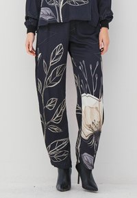 Dark pants with large beige and gray floral patterns worn with black pointed high-heeled ankle boots, hands relaxed by sides.