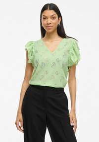 Top verde chiaro a maniche corte con scollo a V e tessuto texturizzato caratterizzato da un motivo a spirale. Abbinato a pantaloni neri.