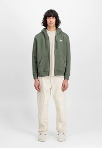 Olivovo zelená mikina na zips so sťahovacou šnúrkou, prednými vreckami a bielym logom; kombinovaná s krémovými nohavicami a svetlými topánkami.