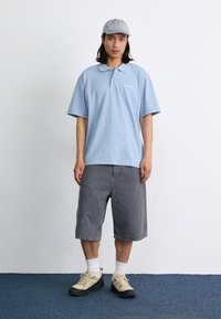 Polo shirt bleu clair avec col, short gris jusqu'aux genoux, chaussettes blanches et baskets beiges avec semelles noires. Complété par une casquette rayée.