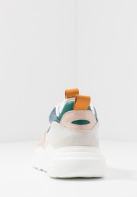 Vue arrière d'une basket multicolore avec une semelle blanche, des détails en daim beige, du cuir vert et des languettes orange sur fond blanc.