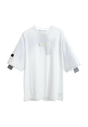 Joven de pie mirando hacia adelante con una camiseta blanca extragrande y pantalones negros con piernas inferiores desmontables y rayas laterales grises.