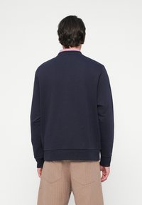 Moški, oblečen v mornarsko modro majico s kapuco čez roza ovratnik in bež hlače, prikazan zadaj na belem ozadju.