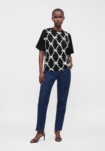Femme debout portant une chemise noire avec des motifs de nœuds blancs, un jean bleu et des sandales à talons noirs sur un fond clair uni.