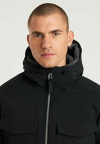 Chaqueta negra con cremallera frontal, cuello alto y capucha. Cuenta con dos bolsillos frontales y una textura de tela suave. Detalle de cremallera plateada.