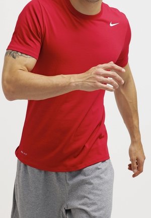 Mężczyzna w czerwonej koszulce Nike Dri-FIT i szarych krótkich spodenkach w pozycji biegowej z tatuażem na lewej górnej części ramienia.