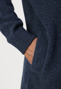 Pulover tricotat texturat de culoare bleumarin, cu o croială lejeră, manșete cu nervuri și un buzunar pe lateral. Material moale și pufoasă vizibil în prim-plan.