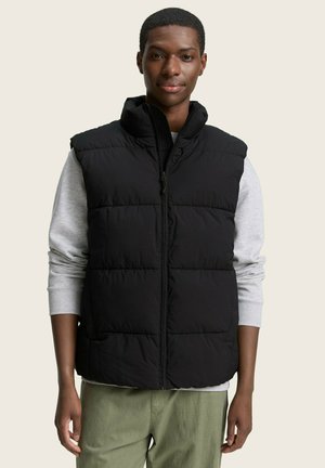 Chaleco acolchado negro con cuello alto y cremallera frontal, puesto sobre un suéter gris claro. El modelo se encuentra frente a un fondo neutro.