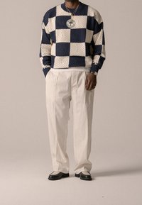 Marineblauer und weiß karierter Strickpullover mit entspannter Passform, kombiniert mit lockeren weißen Hosen und schwarzen Schuhen mit Mesh-Details.