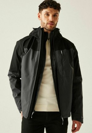 Młody mężczyzna ubrany w czarno-szarą kurtkę na zamek z beżowym swetrem, stojący na tle jasnego, gładkiego tła.