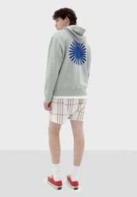 Grijze hoodie met een blauwe zonnestraalafbeelding op de achterkant, gecombineerd met geruite shorts in wit, rood en blauw, en rode schoenen.